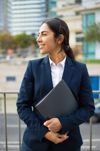 Content businesswoman holding folder. Smiling young businesswoman in formal wear holding papers and looking aside on street. Professional occupation concept